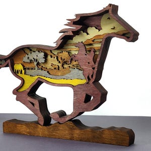 Op de afbeelding: Een meerlaagse houten sculptuur van een rennend paard met een cowboy. De sculptuur gebruikt tinten bruin, geel en grijs hout. Het paard staat tegen een landschap met bomen, een huis en een zonsondergang.