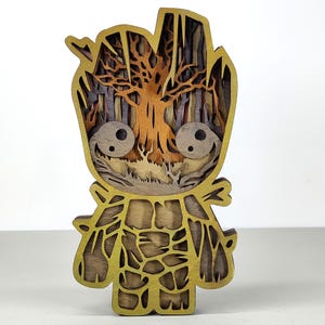 以下が含まれることがあります： 木をモチーフにしたデザインのキャラクターの、多層構造の木製彫刻。明るい茶色と金色の配色で、内部には木と森の風景が描かれています。キャラクターは大きく丸い目を持っています。