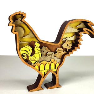 May include: A layered wooden rooster sculpture with a rural landscape scene inside. The rooster is primarily brown and yellow, with a detailed cutout design. The interior features a yellow sun, clouds, a fence, and small houses, creating a three-dimensional effect.
