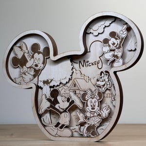 May include: Wooden Mickey Mouse-shaped wall art featuring layered cutouts of Mickey, Minnie, and other Disney characters. The word "Mickey" is written in script. The art piece is made of light-colored wood and has a three-dimensional effect.