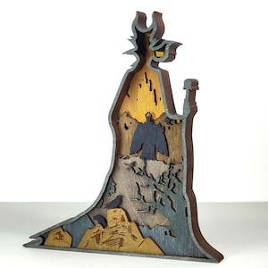Puede incluir: Escultura de madera multicapa de un personaje con un sombrero alto y puntiagudo y una larga t&uacute;nica. La escultura presenta tonos de marr&oacute;n, gris y amarillo, con intrincados detalles recortados que representan una escena de paisaje. El dise&ntilde;o general es una representaci&oacute;n art&iacute;stica estilizada.