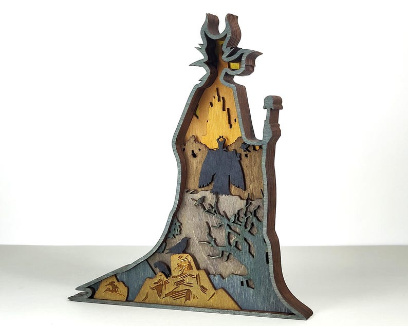 Puede incluir: Escultura de madera multicapa de un personaje con un sombrero alto y puntiagudo y una larga t&uacute;nica. La escultura presenta tonos de marr&oacute;n, gris y amarillo, con intrincados detalles recortados que representan una escena de paisaje. El dise&ntilde;o general es una representaci&oacute;n art&iacute;stica estilizada.