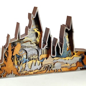 Puede incluir: Escultura de madera multicapa que representa un paisaje de fantasía con un castillo, montañas y un puente. La escultura presenta varios tonos de marrón, gris y azul, con un fénix dorado. La pieza mide aproximadamente 25 cm de ancho.