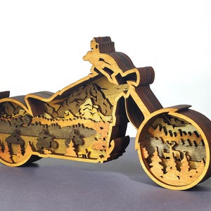 May include: A wooden motorcycle sculpture with a layered design. The sculpture features a detailed landscape scene with mountains, trees, and riders. The wood has a natural finish with varying shades of brown and tan.