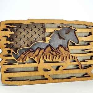 May include: A wooden layered art piece depicting an American flag with a horse silhouette, mountains, and birds. The flag's stars are cut out, and the horse and mountains are layered for depth. The piece is made of natural wood tones.