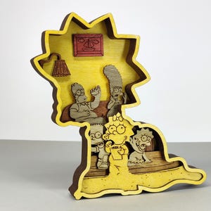 May include: A layered wooden art piece featuring the Simpson family. The artwork is in the shape of Marge Simpson's head, with the family inside a living room scene. The piece is primarily yellow, brown, and tan.