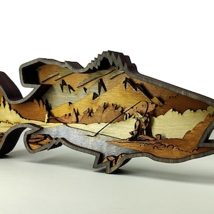 May include: A layered wooden fish sculpture depicting a fishing scene. The sculpture has a layered design with a mountain landscape and a fisherman. The fish is made of various shades of brown wood, with a layered effect.