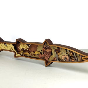 Könnte beinhalten: Ein mehrschichtiges Holzkunstwerk in Form eines Fisches. Das Kunstwerk zeigt Szenen eines springenden Fisches, eines Fischers und einer Landschaft. Die fischförmige Kunst besteht aus verschiedenen Braun- und Beigetönen.