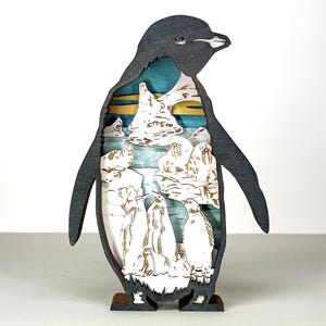 May include: A wooden penguin-shaped art piece with a layered design. The outer layer is dark gray, while the interior depicts a snowy landscape with penguins, icebergs, and a blue and gold sky. The base is brown.