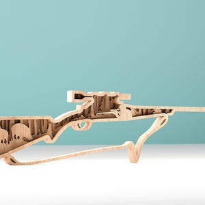 Könnte beinhalten: Eine Holzskulptur in Gewehrform mit einer geschnitzten Waldszene im Inneren. Die hellfarbene Holzschnitzerei zeigt einen Bären, Bäume und einen Jäger. Das Gewehr wird von einem geschwungenen Ständer getragen, was es zu einem einzigartigen Dekorationsartikel macht.