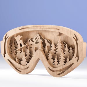 May include: Wooden ski goggles-shaped wall art. The art features a skier, mountains, and trees, all cut from wood. The goggles are light brown and have a layered design, creating a 3D effect. The background is a light blue.
