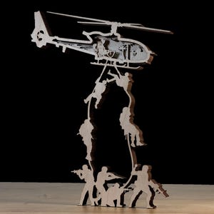May include: A wooden sculpture depicting a helicopter with soldiers rappelling down ropes. The sculpture features detailed cutouts of the helicopter's interior and the soldiers, with a light brown color and a dark background.
