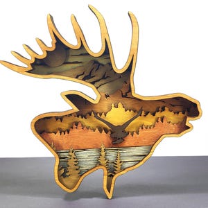 May include: Wooden moose-shaped wall art with a layered design showing a sunset over a lake and mountains. The artwork features a flying bird silhouette and detailed trees, with natural wood tones and a warm colour palette.