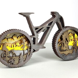 May include: A wooden mountain bike sculpture with intricate layered design. The wheels feature a landscape scene with cyclists, trees, and mountains. The bike is brown with a gray finish, creating a three-dimensional effect. A decorative item for bike enthusiasts.