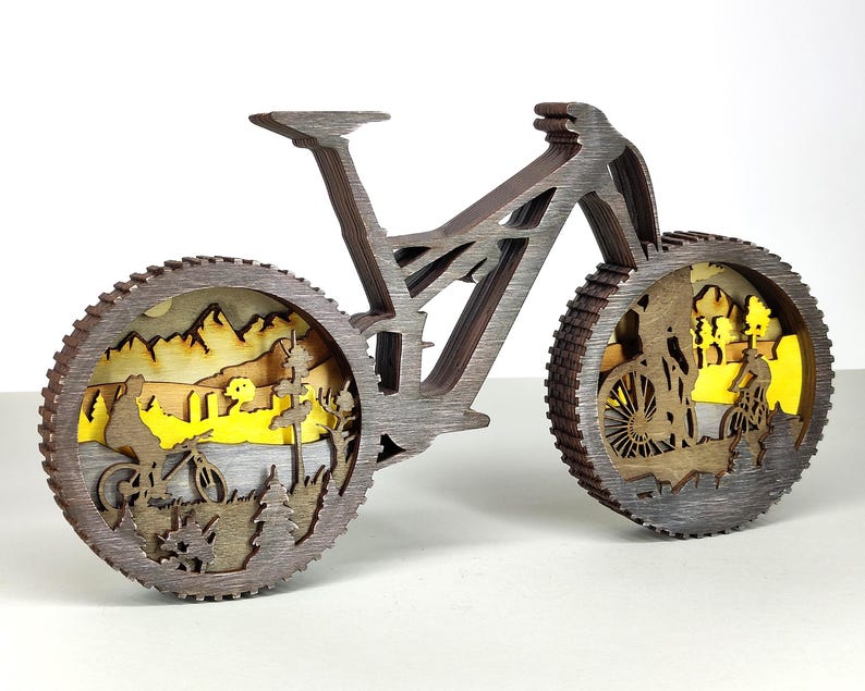 May include: A wooden mountain bike sculpture with intricate layered design. The wheels feature a landscape scene with cyclists, trees, and mountains. The bike is brown with a gray finish, creating a three-dimensional effect. A decorative item for bike enthusiasts.