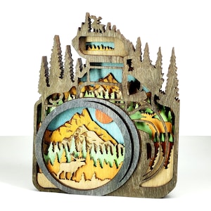 May include: A wooden layered art piece in the shape of a camera, featuring a mountain landscape scene. The design incorporates trees, a sun, and a deer, with a color palette of brown, blue, yellow, and green.