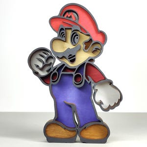 Può includere: Una figura in legno a strati del personaggio del videogioco Mario. La figura presenta un cappello rosso, una tuta blu e un viso color beige. La figura è realizzata con più strati di legno, creando un effetto 3D. La figura è in piedi e ha un'espressione amichevole.