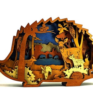 Könnte beinhalten: Ein mehrschichtiges Holzkunstwerk in Form eines Igels, das eine Waldszene darstellt. Das Kunstwerk zeigt Hirsche, eine Eule, ein Eichhörnchen und andere Waldtiere in einem mehrschichtigen Design mit Braun-, Blau- und Gelbtönen.