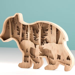 May include: A wooden bear-shaped decorative piece with a layered design. The interior features a forest scene with trees and other bears. The piece is made of light-colored wood and stands upright.
