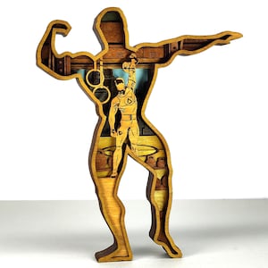 May include: A layered wooden sculpture of a bodybuilder. The sculpture has a detailed interior scene of a weightlifter, gym rings, and gym equipment. The wood has a natural, warm tone with visible grain.