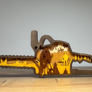 May include: A wooden chainsaw-shaped art piece with a layered design. The piece features a dark brown exterior and a yellow and brown interior, depicting a forest scene with figures and deer.