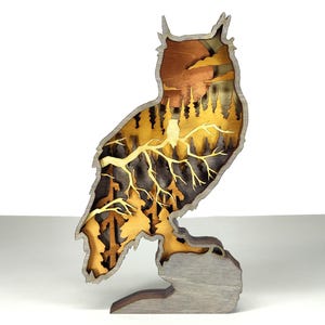 Può includere: Una scultura di gufo in legno con un design a strati. Il gufo è appollaiato su una base, con una scena di foresta visibile all'interno della sua silhouette. La scultura presenta sfumature di marrone, oro e grigio, creando un effetto tridimensionale.