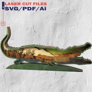 May include: A layered wooden alligator sculpture with a detailed interior landscape. The artwork features shades of green, brown, and tan, with the text "LASER CUT FILES SVG/PDF/Ai" in the upper left corner.