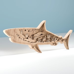 May include: A light brown wooden shark sculpture, intricately layered to show an underwater scene. The design includes a diver, coral, and marine life. The shark is displayed on a white surface with a blue background.