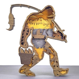 May include: A wooden Bigfoot-shaped art piece with a fishing theme. The layered design features a Bigfoot silhouette holding a fishing rod, with a lake and mountain scene inside. The color palette includes shades of brown, yellow, and blue.