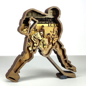 May include: A wooden hockey player sculpture in a layered design. The sculpture depicts a hockey player in action, with a detailed interior scene of a hockey game. The sculpture is made of light and dark brown wood.