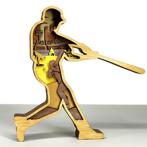 May include: A multi-layered wooden baseball player figurine in a batting stance. The interior layers depict a baseball field and cityscape, with shades of yellow, brown, and tan. The figurine is a decorative item, perfect for sports enthusiasts.