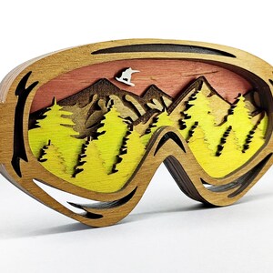 May include: Wooden art piece in the shape of ski goggles, featuring a layered design of a mountain scene. The scene includes a skier, mountains, and yellow and green trees, set against a sunset sky. The frame is a natural wood color.