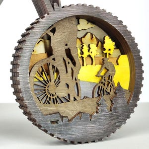 May include: A layered wooden bicycle-themed decoration. The design features a cyclist riding a mountain bike, with a yellow and brown landscape background. The outer frame is a gear shape with a gray finish.