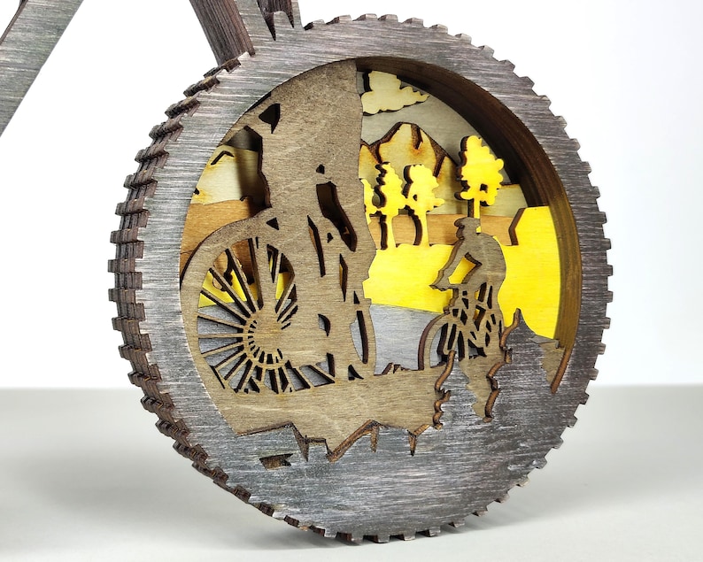 May include: A layered wooden bicycle-themed decoration. The design features a cyclist riding a mountain bike, with a yellow and brown landscape background. The outer frame is a gear shape with a gray finish.