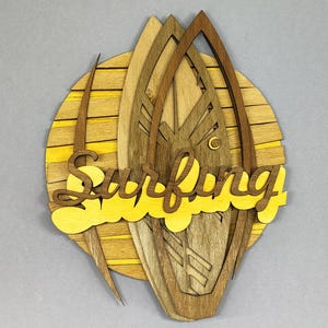 May include: Wooden wall art featuring a layered design of surfboards and a sun. The word "Surfing" is cut out in yellow. The art piece is made of various shades of brown and yellow wood, creating a dimensional effect.