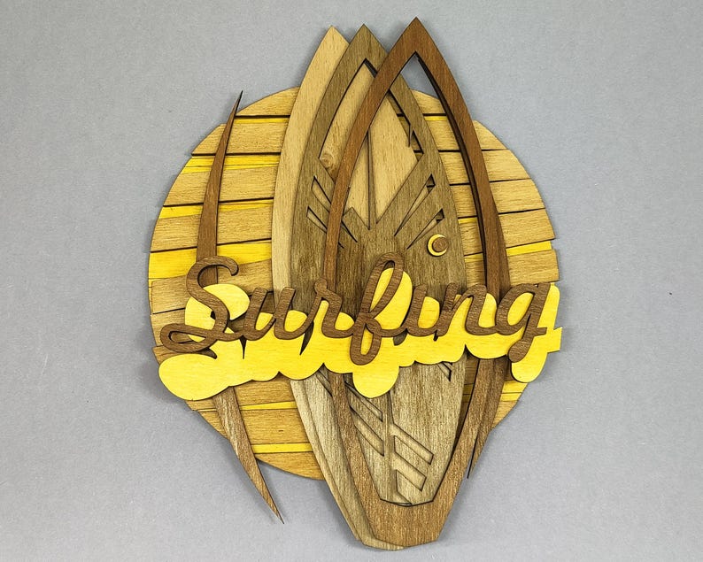 May include: Wooden wall art featuring a layered design of surfboards and a sun. The word "Surfing" is cut out in yellow. The art piece is made of various shades of brown and yellow wood, creating a dimensional effect.