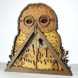 May include: A wooden owl sculpture with layered details. The owl wears round glasses and has a detailed feather pattern. Inside a triangular frame, a castle and forest scene is visible. The sculpture is made of various shades of brown wood.