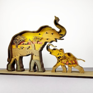 May include: A layered wooden artwork showing an adult elephant and a calf. The elephants' interiors display a savanna scene with trees, birds, and a sunset. The piece is mounted on a rectangular wooden base.