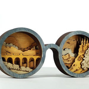 May include: A wooden decorative piece shaped like eyeglasses, featuring layered laser-cut scenes. One lens depicts a train on a bridge, the other a castle. The frames are a muted blue, and the interior scenes are natural wood tones.