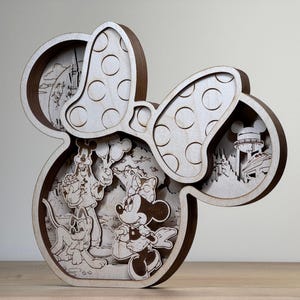 May include: Wooden layered art piece in the shape of Minnie Mouse's head with a bow. The artwork features scenes with Mickey, Minnie, Pluto, and park attractions. The bow has white circles on it. The piece is made of light and dark brown wood.