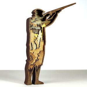 Peut inclure: Sculpture en bois multicouche représentant une personne visant avec un fusil. La figurine est marron avec un intérieur découpé révélant un paysage avec des arbres et une figurine jaune. La sculpture est sur une surface blanche.