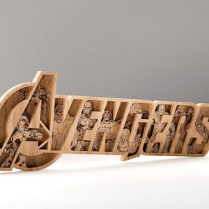 Puede incluir: Letrero de madera de los Vengadores con figuras de superhéroes grabadas dentro de las letras. El letrero está hecho de madera de color claro y tiene un diseño tridimensional. El logotipo de los Vengadores se muestra de forma destacada.