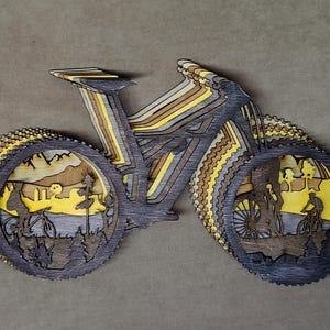 May include: A layered wooden art piece depicting a mountain bike. The artwork features a multi-layered design with shades of gray, brown, and yellow. The wheels showcase scenes of cyclists in a natural landscape.