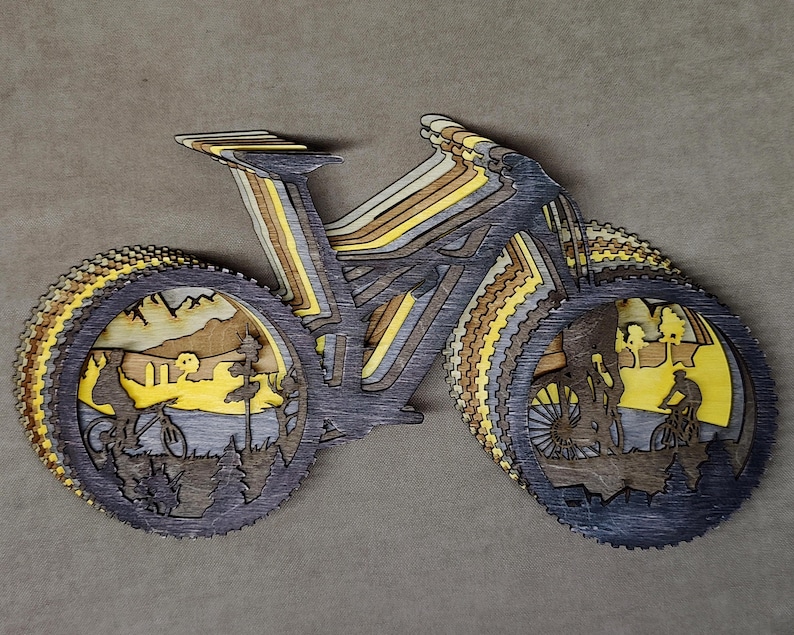 May include: A layered wooden art piece depicting a mountain bike. The artwork features a multi-layered design with shades of gray, brown, and yellow. The wheels showcase scenes of cyclists in a natural landscape.