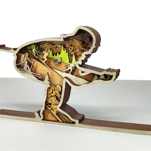 May include: A wooden sculpture of a skier in motion, with a detailed interior scene of skiers on a mountain slope. The sculpture is made of layered wood in shades of brown, tan, and green, with a white background.