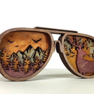 Può includere: Occhiali da sole da aviatore in legno con un design a strati raffigurante un paesaggio montano con uccelli e un cervo in una foresta. Le montature sono marroni e le scene interne presentano tonalità arancioni, viola e verdi.