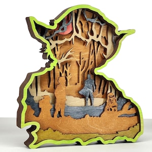 May include: A layered wooden art piece in the shape of Yoda from Star Wars. The artwork features a green outline, with various shades of brown, gray, and tan depicting a forest scene with characters. The piece is designed to stand upright.