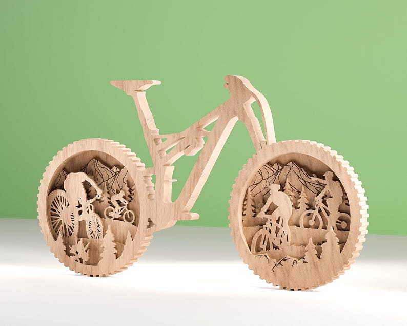 May include: A wooden mountain bike sculpture with intricate cutouts. The wheels feature scenes of cyclists riding through a mountain landscape with trees and peaks. The bike is made of light-colored wood and stands against a green background.