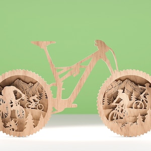 May include: Wooden mountain bike art piece. The artwork features a detailed silhouette of a mountain bike with riders, trees, and mountains. The design is made of light-colored wood and is set against a green background.