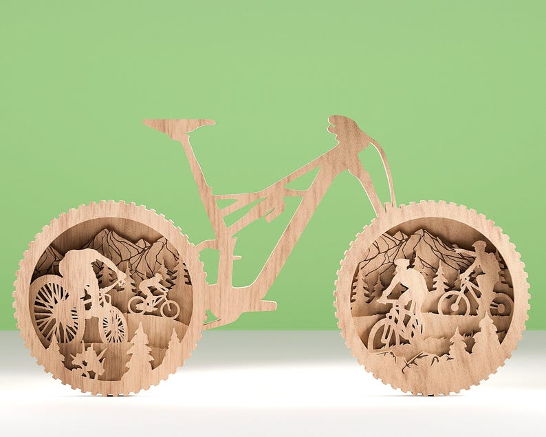 May include: Wooden mountain bike art piece. The artwork features a detailed silhouette of a mountain bike with riders, trees, and mountains. The design is made of light-colored wood and is set against a green background.